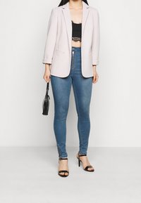 Blazer rosa claro con mangas remangadas, top corto negro con borde de encaje, jeans pitillo azules con cremalleras y tacones negros con tiras.