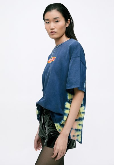 Modrá tričko s tie-dye vzorem, které má živou grafickou nálepku, krátké rukávy a volný střih. K tričku jsou sladěny lesklé černé šortky.