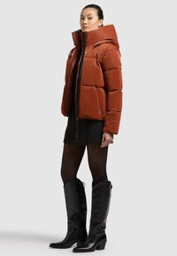 Ruda pūkinė striukė su aukštu apykakle, užtrauktuku ir dviem priekinėmis kišenėmis. Stilizuota su juoda suknele ir juodais ilgaauliais batais.