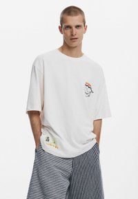 Tricou alb din bumbac, cu o croială lejeră, având o mică imprimare grafică pe piept și un design colorat cu palmieri la margine.