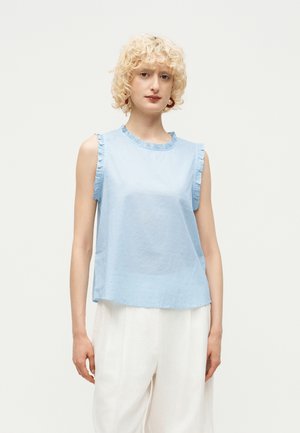 Γυναίκα με ξανθά σγουρά μαλλιά που φοράει αμάνικο ανοιχτό μπλε τοπ με βολάν και λευκά παντελόνια φαρδιάς γραμμής σε λευκό φόντο.