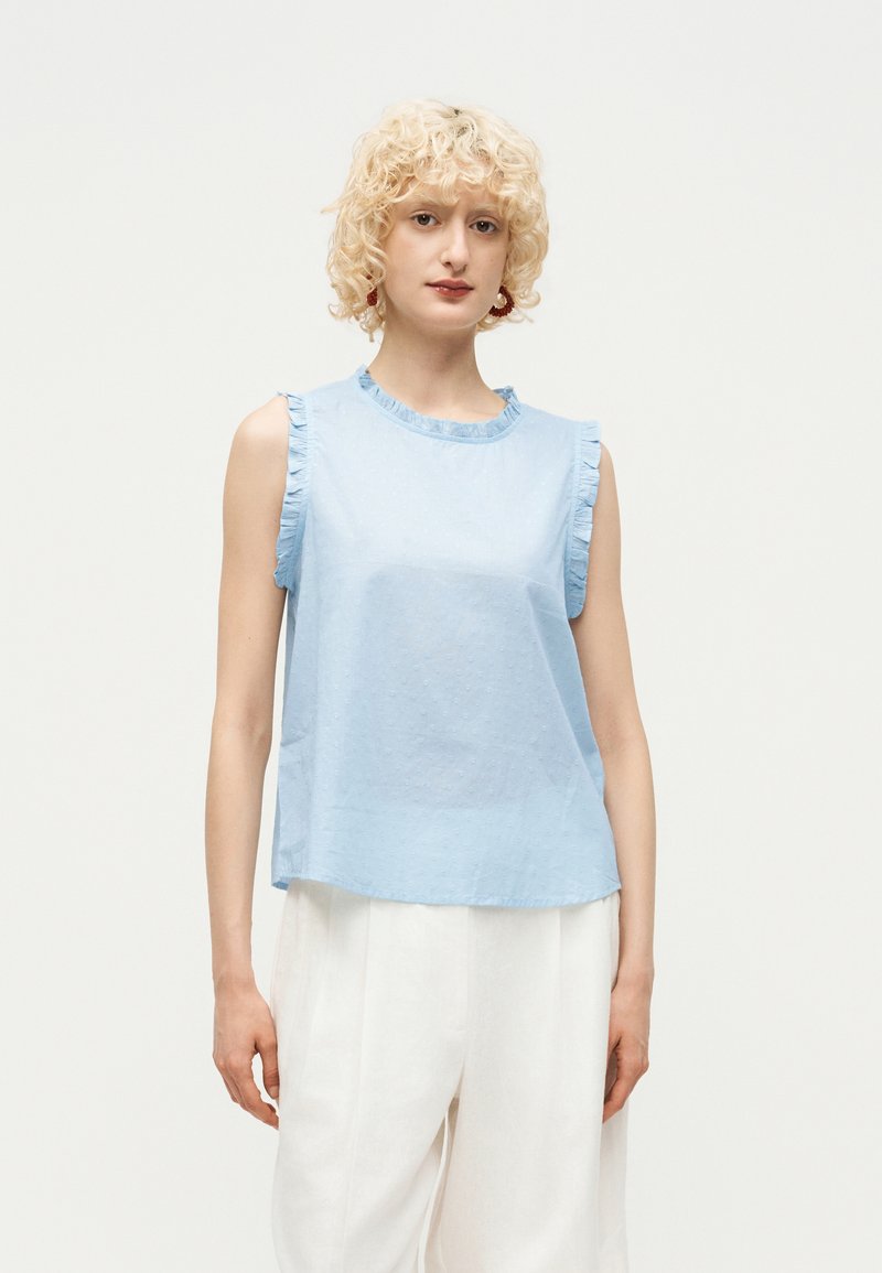 Γυναίκα με ξανθά σγουρά μαλλιά που φοράει αμάνικο ανοιχτό μπλε τοπ με βολάν και λευκά παντελόνια φαρδιάς γραμμής σε λευκό φόντο.