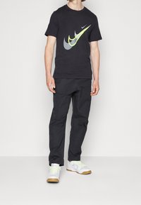 Černé tričko Nike s velkým neonově žlutým a šedým logem, doplněné černými kalhotami a bílými teniskami s gumovou podrážkou a neonovými detaily.