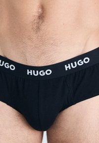 Schwarze Unterwäsche aus einem weichen, dehnbaren Stoff mit einem breiten elastischen Taillenbund, der das Wort "HUGO" in weißer Schrift zeigt.