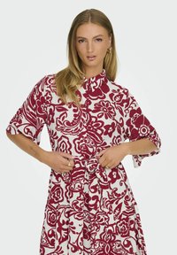 Abito a fiori rosso e bianco con colletto, maniche corte e cintura in vita. Il tessuto appare strutturato con un design leggero e fluido.