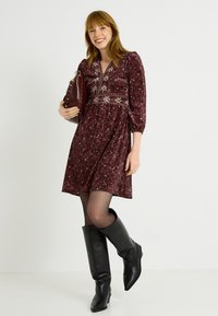 Vestido floral burdeos con cintura fruncida, mangas largas abullonadas y escote en V, combinado con botas negras hasta la rodilla y un bolso.