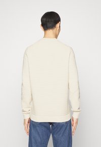 Homme de dos portant un pull côtelé crème et un jean bleu sur un fond clair uni.