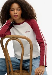 Langarm baseball t-shirt met een witte boddy en burgundy raglan mouwen, voorzien van witte strepen; "Superdry" logo geborduurd op de borst.