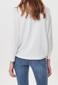 Donna dai lunghi capelli castani, che indossa un maglione bianco a coste e jeans blu, in piedi di spalle contro uno sfondo semplice.