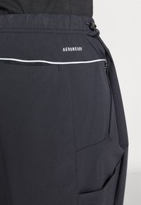 Crne sportske hlače s podesivim strukom, s bijelim logotipom i reflektirajućim detaljima. Tkanina izgleda rastezljivo i glatko.