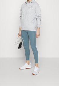Sweat à capuche gris avec une poche avant, associé à des leggings bleu sarcelle et des baskets blanches. La tenue comprend une corde à sauter tenue dans une main.
