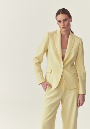 Vrouw met lichtbruin haar in een geel op maat gemaakt pak met een blazer met één knoop en bijpassende broek, poserend tegen een effen gebroken witte achtergrond.