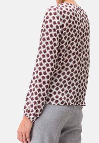 Femme portant un blouse à manches longues avec un motif géométrique maroon, blanc et noir, associée à un pantalon gris clair, vue de dos.