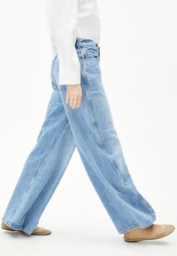 Helle blaue Denim-Breite-Hosen mit hoher Taille, rohen Säumen und auffälligen Seitennähten, kombiniert mit beigen Slippern.