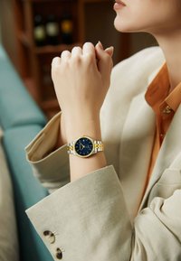 Reloj de metal dorado y plateado con una esfera de color azul marino, que presenta una caja redonda y una correa de eslabones, llevado en una mano con uñas arregladas.