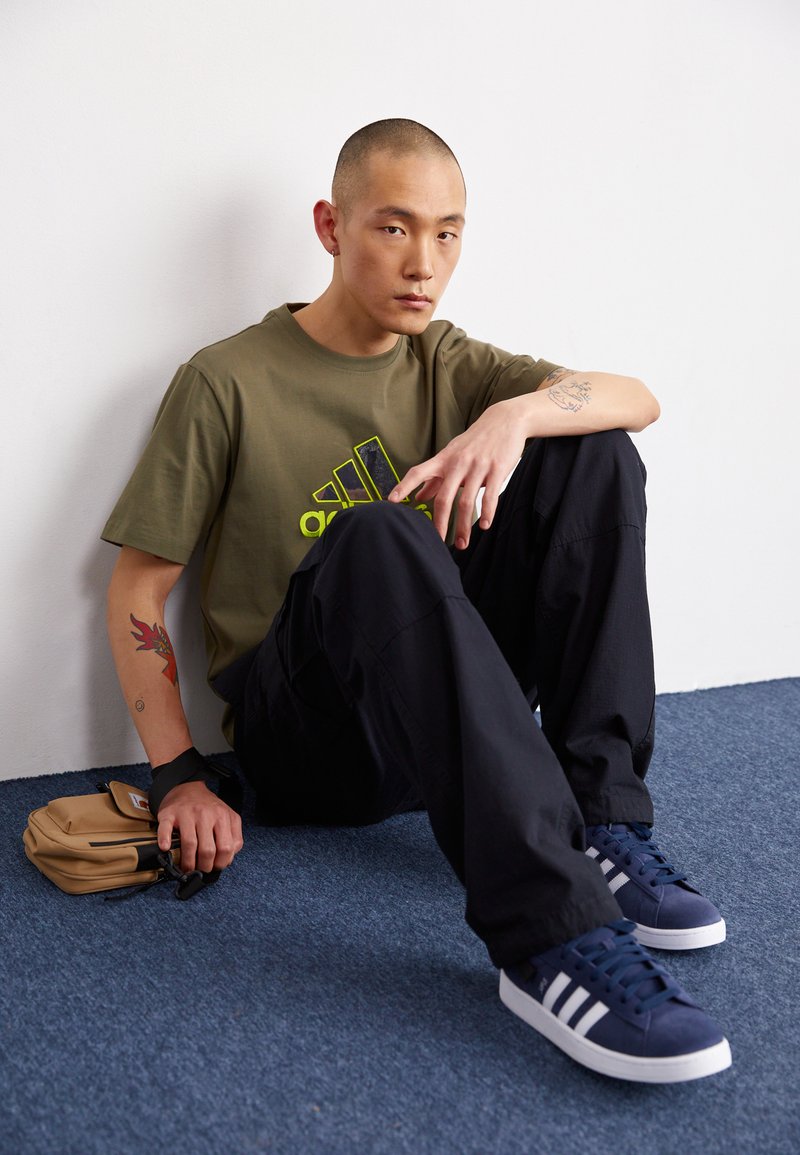 Olijfgroene t-shirt met zwart logo, zwarte baggy broek en donkerblauwe sneakers met witte strepen. Gladde texturen, zittende houding op een blauw tapijt.