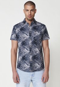 Camisa de manga corta en azul marino con un patrón de hojas de palma blancas, diseño de botonadura frontal y un sutil parche con el logo. Fabricada en tela ligera.