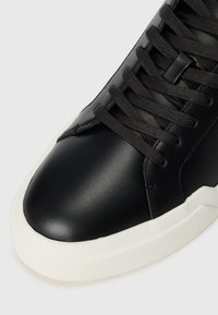 Sneaker din piele neagră cu vârful rotunjit, șireturi negre și talpă din cauciuc alb. Suprafață netedă cu detalii de coasere minime.