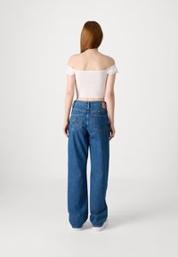 Široké modré džíny s výšivkou květin na zadních kapsách, kombinované s bílým topem s odhalenými rameny. Hladká textura, ležérní design.