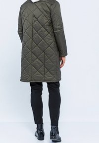 Grøn quiltet frakke med lige snit, der har hætte og lange ærmer. Båret over sorte bukser og ankelstøvler.