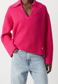 Różowy sweter prążkowany z dekoltem w kształcie litery V i dużym kołnierzem. Posiada długie rękawy, luźny krój oraz małe złote logo w kształcie serca po boku.