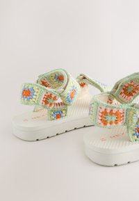Sandales style crochet avec motifs floraux multicolores dans des tons pastel, avec une bride vert clair et des semelles blanches texturées.