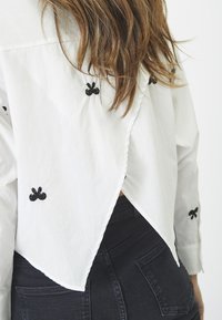 Camicia bianca con bottoni e fiocchi neri ricamati, caratterizzata da un orlo posteriore profondo e maniche lunghe, abbinata a jeans scuri.