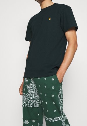 Person iført en sort t-shirt med korte ærmer med et lille gult logo og grønne bukser med hvide paisley- og geometriske mønstre.