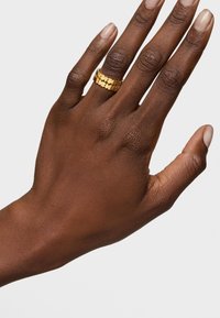 Złoty pierścionek na dłoni, z wyjątkowym wyciętym wzorem. Gładka faktura i polerowane wykończenie kontrastujące z ciemnym odcieniem skóry.