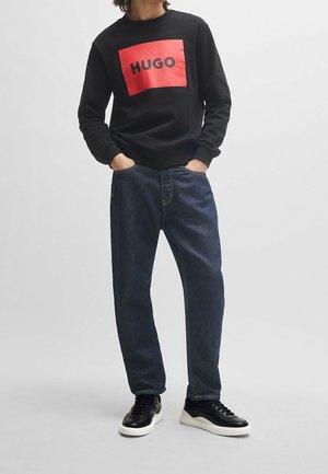 Sweatshirt noir avec un patch rectangulaire rouge bien visible portant le mot "HUGO" en noir ; associé à un jean en denim foncé ample et des baskets noires.