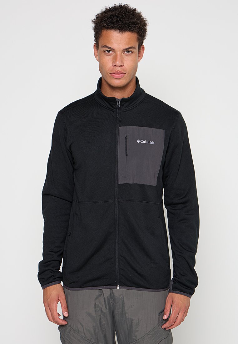 Veste en polaire noire avec une poche sur la poitrine grise ; elle possède une fermeture éclair complète et un col haut. Texture douce et manches longues, idéale pour le superposition.