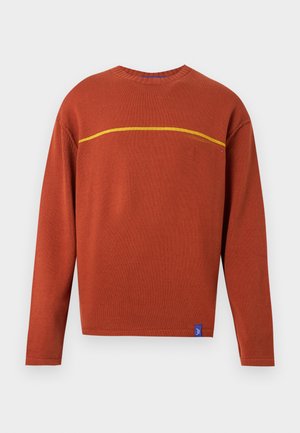 Muškarac u puloveru boje hrđe s žutom prugom, crnim širokim trapericama, crnim cipelama i smeđom kapom stoji s rukama iza leđa.