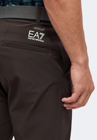 Gros plan sur la taille et la main d’une personne portant un pantalon marron foncé avec le logo Emporio Armani EA7 Ventus et une ceinture noire.