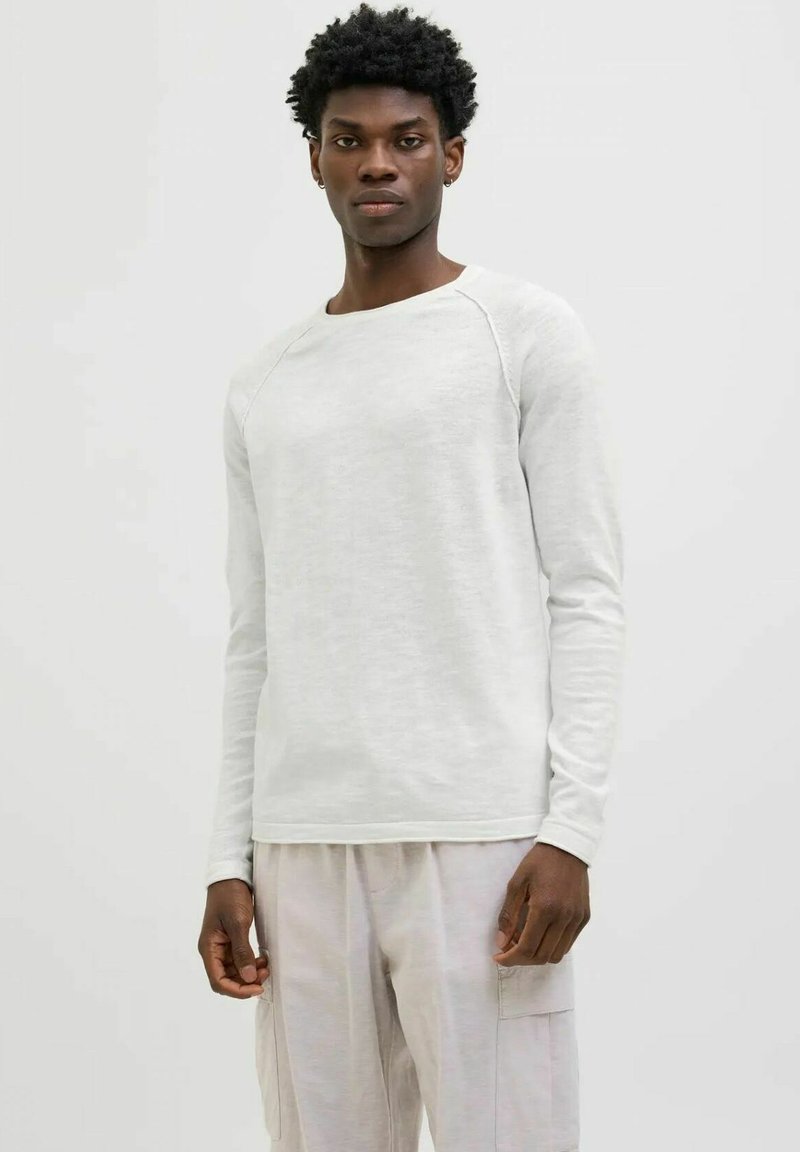 Camicia a maniche lunghe grigia chiara in un tessuto morbido e texturizzato, con scollatura a girocollo e maniche raglan, abbinata a pantaloni larghi di colore chiaro.
