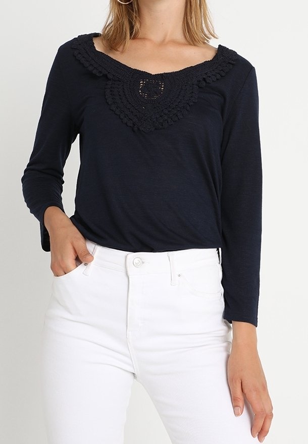 Navyblaues Longsleeve-Oberteil mit Häkel-Ausschnitt und strukturierten Akzenten, kombiniert mit weißen, hochgeschnittenen Hosen.