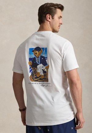 Homme portant un t-shirt blanc avec un motif d’un ours assis en tenue nautique, un short bleu marine et une montre-bracelet à son poignet gauche.