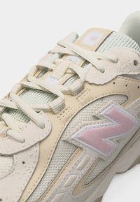 Beige ja koorevärvi New Balance tossu lähipilt, millel on roosa logo ja valged paelad võrgust ja nubuknahast tekstuuriga pealispinnal.