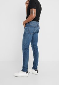 Vaqueros de denim azul oscuro con corte slim, diseño de cinco bolsillos y costuras en contraste, combinados con zapatillas blancas y una camisa negra ajustada.