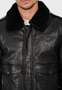 Černá kožená bunda s ovčím límcem, vybavená předním zipem, patentkami a dvěma předními kapsami. Vystouplý detail s křídly.