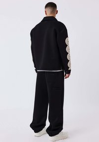Zwarte oversized jas met crèmekleurige scalloped borduursels op de mouwen, gecombineerd met zwarte wijde broek en witte sneakers. Glad stoftextuur.