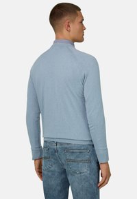 Jeune homme de profil portant un pull bleu clair par-dessus une chemise à col et un jean bleu, sur un fond uni.