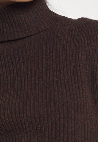 Brauner, gerippter Rollkragenpullover mit strukturiertem Finish, der einen hohen Kragen und eine taillierte Passform aufweist. Warme, erdige Töne betonen das Material.