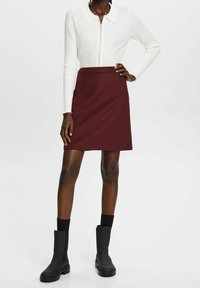 Femme portant un pull blanc côtelé à fermeture éclair, une jupe bordeaux jusqu'aux genoux, des chaussettes noires et des bottines noires, debout avec une main sur la hanche.