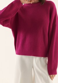Magenta Strickpullover mit lockerer Passform, gerippter Textur und weit geschnittenen Ärmeln. Kombiniert mit weißen weit geschnittenen Hosen, die die Länge des Pullovers zur Geltung bringen.