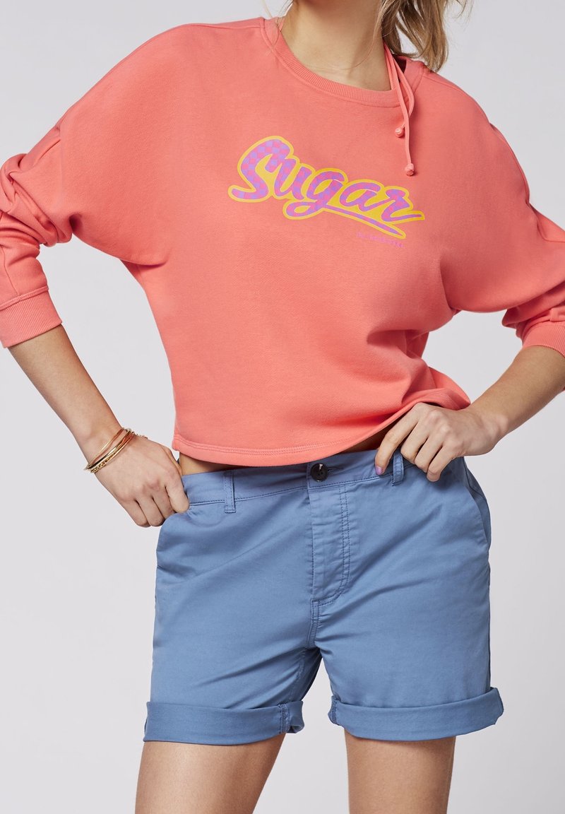 Personne portant un sweat-shirt court corail avec le texte "Sugar" et un short bleu clair roulé, les mains posées sur les hanches.