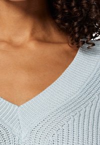 Nahaufnahme eines Halses und oberen Brustbereichs einer Person, die einen hellblauen, gestrickten V-Ausschnitt-Pullover trägt, mit lockigem, dunklem Haar, das auf einer Seite sichtbar ist.