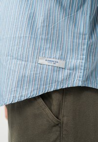 Μπλε ανοιχτό και λευκό ριγέ πουκάμισο με ετικέτα «ELEMENT CO. SINCE 1992» πάνω από την τσέπη του χακί παντελονιού.