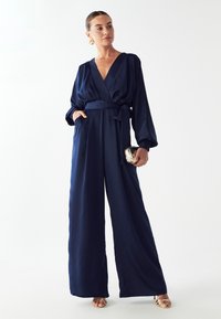 Navy blauwe jumpsuit met een diepe V-hals, brede mouwen en een gestrikt taille. Gladde, satijnachtige textuur met zijzakken en wijde pijpen.