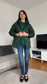 Quiltet grønn jakke med glidelås og hette, brukt over en grønn skjorte, kombinert med blå jeans og svarte flate sko, vist innendørs.