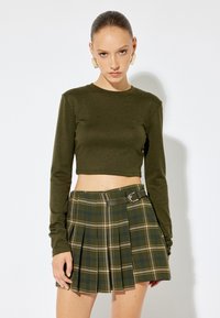 Olivgrünes, cropped Langarmshirt kombiniert mit einem gefalteten Karorock, der grüne, blaue und gelbe Akzente hat, mit einem Gürtel gesichert.